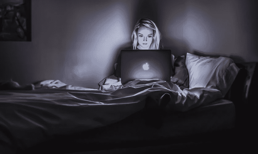 A woman sits in bed, eyes glued to her computer.
