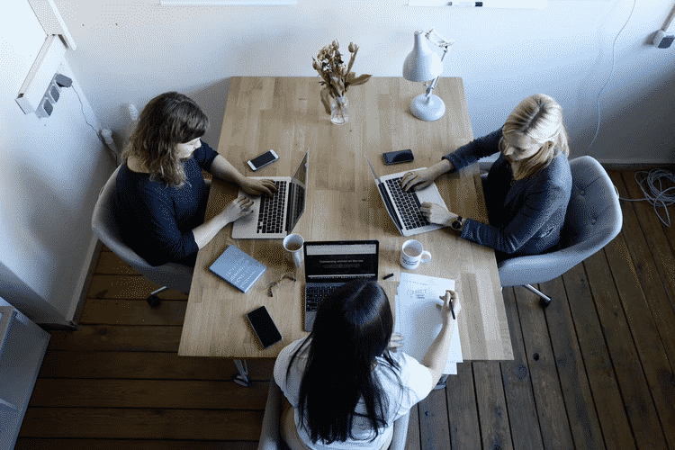 Three women sit at a table with their laptops and conduct research